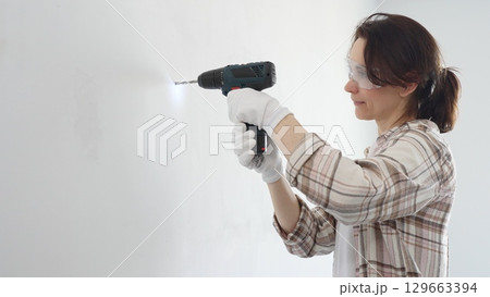 Middle aged female professional worker wearing protective gear and gloves, is drilling hole in white wall during home improvement project, focused on precise home renovation work 129663394