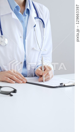 Unknown female doctor completing healthcare paperwork, clipboard positioned near laptop and stethoscope in well lit clinical workspace by window. Medicine and health care Unknown female doctor completing healthcare paperwork, clipboard positioned near laptop and stethoscope in well lit clinical workspace by window. Medicine and health care 129663397