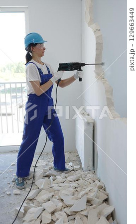 Woman construction worker wearing blue overalls, safety glasses and hardhat is demolishing interior wall with rotary hammer, creating substantial concrete debris pile during demolition process Woman construction worker wearing blue overalls, safety glasses and hardhat is demolishing interior wall with rotary hammer, creating substantial concrete debris pile during demolition process 129663449