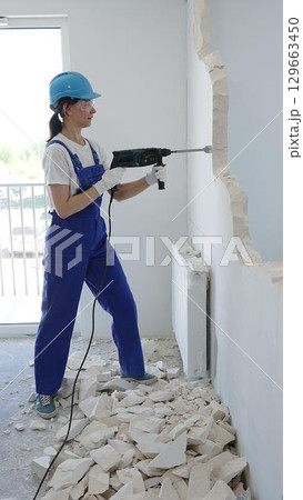 Woman construction worker wearing blue overalls, safety glasses and hardhat is demolishing interior wall with rotary hammer, creating substantial concrete debris pile during demolition process Woman construction worker wearing blue overalls, safety glasses and hardhat is demolishing interior wall with rotary hammer, creating substantial concrete debris pile during demolition process 129663450