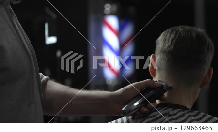 Male professional barber trimming client's hair with electric clipper, vintage illuminated barber pole visible in background, creating contemporary grooming experience Male professional barber trimming client's hair with electric clipper, vintage illuminated barber pole visible in background, creating contemporary grooming experience 129663465