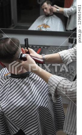 Professional barber grooming client's hair using electric clipper, delivering precise cut with skill in contemporary salon setting 129663481