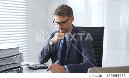 Adult businessman wearing blue suit and tail is concentrating and thinking while working at the desk in a bright, modern office, demonstrating professionalism and efficiency. Business people concept Adult businessman wearing blue suit and tail is concentrating and thinking while working at the desk in a bright, modern office, demonstrating professionalism and efficiency. Business people concept 129663483