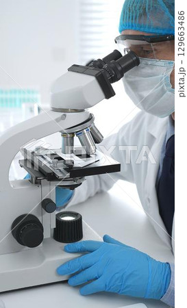 Senior male scientist wearing protective mask, cap and gloves using microscope in laboratory, conducting medical research and analysis. Medicine, health care and science concept Senior male scientist wearing protective mask, cap and gloves using microscope in laboratory, conducting medical research and analysis. Medicine, health care and science concept 129663486