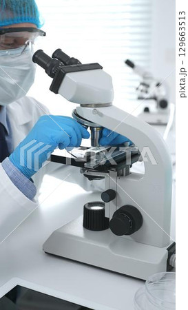 Senior male scientist wearing protective mask, cap and gloves using microscope in laboratory, conducting medical research and analysis. Medicine, health care and science concept Senior male scientist wearing protective mask, cap and gloves using microscope in laboratory, conducting medical research and analysis. Medicine, health care and science concept 129663513