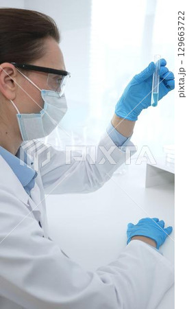 Female researcher examining clear liquid in test tube, sitting near microscopes and lab equipment within sterile scientific workspace. Medicine, healthcare and science concept 129663722