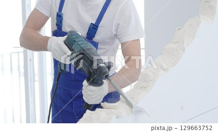 Male construction worker demolishing white wall with rotary hammer drill, wearing blue overalls and protective white gloves, closeup view Male construction worker demolishing white wall with rotary hammer drill, wearing blue overalls and protective white gloves, closeup view 129663752