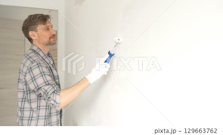 Man professional painter wearing checkered shirt, is rolling white paint on apartment wall, transforming interior space with steady strokes during comprehensive home renovation 129663762