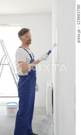 Middle aged man wearing protective white gloves and blue work overalls, applying paint with roller on pristine interior wall during home renovation project, vertical view 129663780