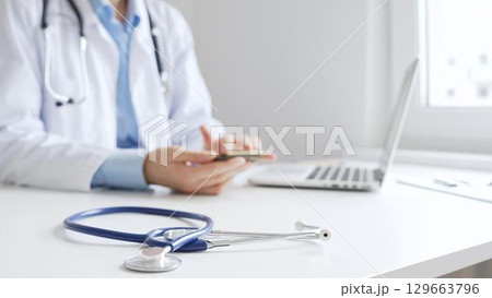 Closeup of blue stethoscope is lying on the table near female doctor checking smartphone representing digital healthcare workflow in well lit clinical workspace. Medicine and health care Closeup of blue stethoscope is lying on the table near female doctor checking smartphone representing digital healthcare workflow in well lit clinical workspace. Medicine and health care 129663796
