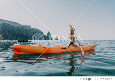 Kayaking Woman Sea Coastal Landscape - A woman enjoys kayaking in calm waters near a scenic coastline with rocky cliffs on a sunny day. 129664681