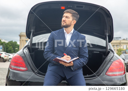 Businessman sitting on car trunk writing notes in a city setting during a cloudy day 129665180