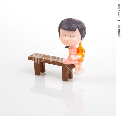Miniature girl sitting on a wooden bench holding a small orange bunny toy in a bright setting 129665186