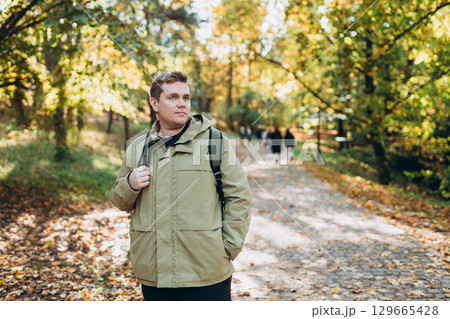 Calm happy 30s Man with backpack Relaxing in Autumn Park Outdoors. Traveler. Falling time. Smiling man in casual clothing standing outdoor, web banner 129665428