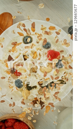 Granola Pouring and Splashing into the White Bowl with Milk. Served on Kitchen Table. Concept of Flying Food. Overhead View. Granola Pouring and Splashing into the White Bowl with Milk. Served on Kitchen Table. Concept of Flying Food. Overhead View. 129665677