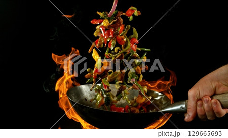 Closeup of chef throwing asian vegetable with meat mix from wok pan in fire. Fresh asian food preparation on black background. Closeup of chef throwing asian vegetable with meat mix from wok pan in fire. Fresh asian food preparation on black background. 129665693