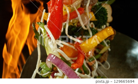 Closeup of chef throwing asian vegetable with meat mix from wok pan in fire. Fresh asian food preparation on black background. Closeup of chef throwing asian vegetable with meat mix from wok pan in fire. Fresh asian food preparation on black background. 129665694