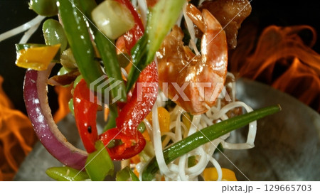 Closeup of chef throwing asian vegetable with meat mix from wok pan in fire. Fresh asian food preparation on black background. Closeup of chef throwing asian vegetable with meat mix from wok pan in fire. Fresh asian food preparation on black background. 129665703