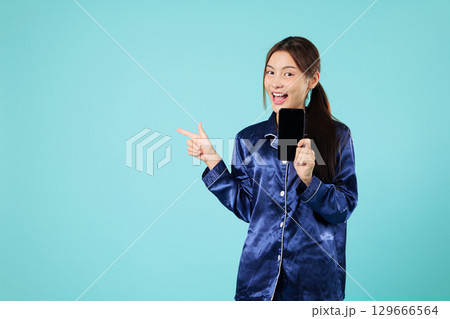 Smiling asian young woman pointing while holding a phone in her pajama, for social media or a sleeping mobile phone app, and looking to the side, headshots isolated studio blue background Smiling asian young woman pointing while holding a phone in her pajama, for social media or a sleeping mobile phone app, and looking to the side, headshots isolated studio blue background 129666564