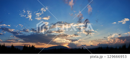 Serene evening sky with hues of blue and soft pink clouds as sun sets. Horizon glows gently, casting light over silhouetted pine trees and rolling hills, creating peaceful and expansive landscape. 129666953