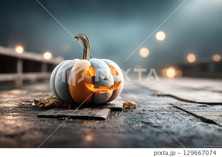 A glowing Halloween pumpkin sits on a wooden surface at night, displaying two distinct colors with mysterious lights in the misty background. 129667074