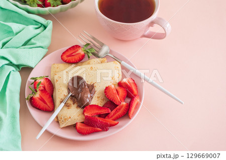 Crepes with chocolate spread and fresh strawberries, served with tea on pink background 129669007