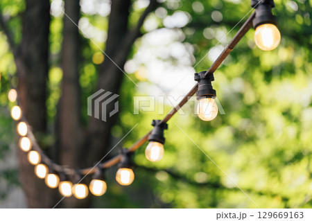 Close up of glowing string lights, street decor in summer garden with blurred green trees background Close up of glowing string lights, street decor in summer garden with blurred green trees background 129669163
