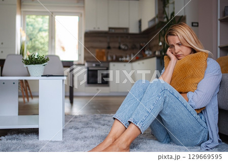 Sad woman sitting on the floor hugging a pillow in her living room 129669595