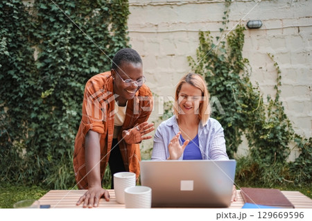 Two businesswomen having a video conference in the garden, waving at the laptop screen Two businesswomen having a video conference in the garden, waving at the laptop screen 129669596