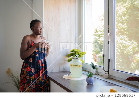 Young woman drinking homemade lemonade and looking out the window in the kitchen Young woman drinking homemade lemonade and looking out the window in the kitchen 129669617