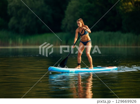 Athletic woman paddleboarding on calm water surrounded by lush greenery and natural scenery 129669926