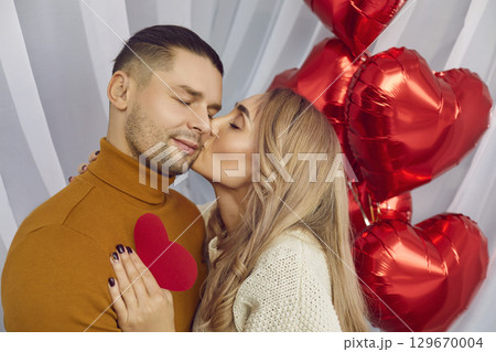 Woman kissing a man and holding red heart in her hand with the background of foil ballons. Woman kissing a man and holding red heart in her hand with the background of foil ballons. 129670004