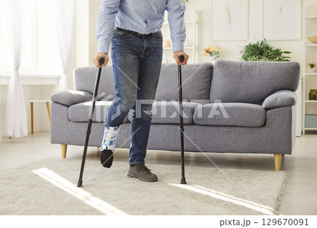 Man with injured leg in plaster cast using crutches and orthopedic boot while recovering at home. 129670091
