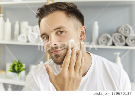 Handsome man smiling looking at camera, mirror, applying cream to cheek, hand gently touching face 129670137