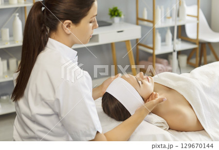 Female beautician making face relaxing massage to a young relaxed woman in beauty spa salon. 129670144