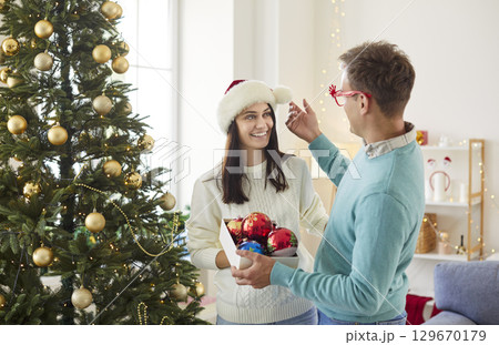 Young couple standing in living room at home decorating Christmas tree together with toy balls Young couple standing in living room at home decorating Christmas tree together with toy balls 129670179