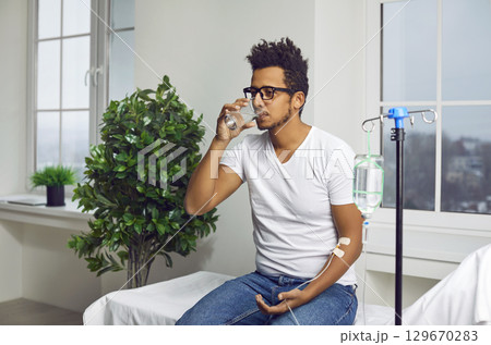 Young man drinks water while receiving vitamin therapy through IV drip line infusion 129670283