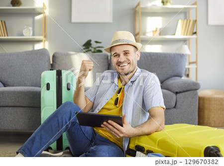 Happy excited young man sitting at home booking tickets online via tablet and looking at camera Happy excited young man sitting at home booking tickets online via tablet and looking at camera 129670382