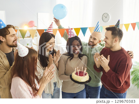 Young happy woman holding her birthday cake with candles standing with friends at home Young happy woman holding her birthday cake with candles standing with friends at home 129670383