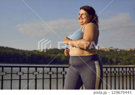 Happy plus size young woman running on a bridge during her jogging workout in summer 129670445