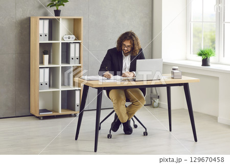 Young stylish business man with long hair working with laptop and writing at the desk in office. Young stylish business man with long hair working with laptop and writing at the desk in office. 129670458
