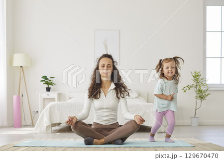 Relaxed mother practicing yoga in lotus pose with closed eyes while playful daughter running around. 129670470
