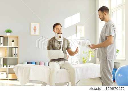 Doctor traumatologist examining man in neck brace with broken arm in medical office in exam room. 129670472