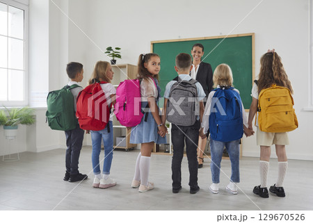Schoolchildren with colorful backpacks on their shoulders talking to teacher in classroom. 129670526