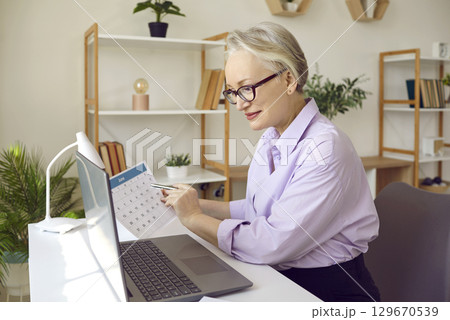 Elegant senior businesswoman checking agenda in office with paper calendar and laptop. Elegant senior businesswoman checking agenda in office with paper calendar and laptop. 129670539