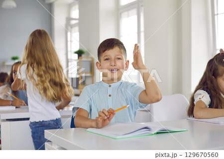 Portrait of small active schoolboy who raises his hand in class to ask question or answer. 129670560