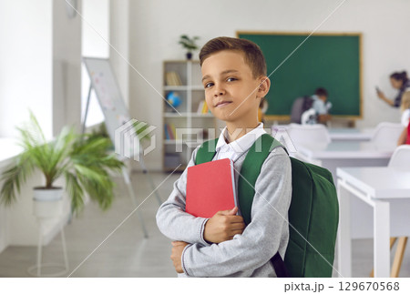 Proud boy first grader with satchel and workbook posing on first day of elementary school in classroom 129670568