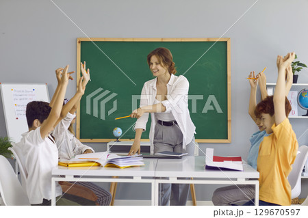 Smiling school teacher asks questions during lesson with group of happy children 129670597