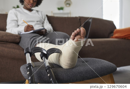 African American woman with broken leg sitting on sofa, with her foot resting on stool African American woman with broken leg sitting on sofa, with her foot resting on stool 129670604