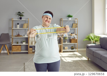 Fat funny woman doing stretching exercises with rubber band in her hands at home. Fat funny woman doing stretching exercises with rubber band in her hands at home. 129670658